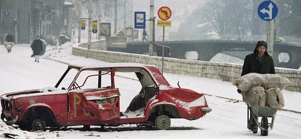 A Sarajevo man pushes his wheelbarrow full of wood, past a car that was destroyed earlier during the siege of Sarajevo on Tuesday, March 9, 1993. The street is normally in open view of Serbian snipers, but light snow or fog sometimes shields pedestrians on the city streets below from snipers. (AP Photo/Michael Stravato)