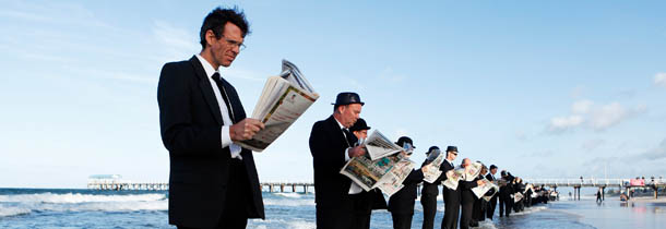 ADELAIDE, AUSTRALIA - JANUARY 08: Volunteers stand and read the morning newspaper while 'waiting for the bus' at Henley Beach on January 8, 2012 in Adelaide, Australia. Surrealist artist, Andrew Baines recruited 100 volunteers for this human installation, meant to illustrate corporate workers enjoying nature rather than waiting in a long queue for a trip to work. (Photo by Morne de Klerk/Getty Images)