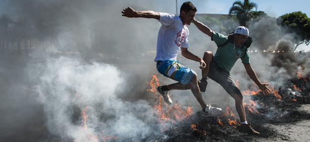 People jump over burnt tyres during a protest blocking the access to Brasilia's Mane Garrincha, one of the six host stadiums for the upcoming Confederations Cup, in Brasilia on June 14, 2013. People demonstrate against the government's policy of the expenditure for the 2014 FIFA World Cup. A police spokesman said the protest was organized by the Homeless Workers Movement (MTST) which is campaigning to reduce Brazil's housing shortage by staging squatters' occupations in abandoned government buildings. Brazil faces Japan Saturday at Mane Garrincha in the opening game of the two-week Confederations Cup, a dry run for next year's World Cup. AFP PHOTO / YASUYOSHI CHIBA (Photo credit should read YASUYOSHI CHIBA/AFP/Getty Images)