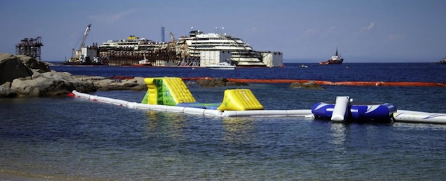 Sunbathers sit on a beach overlooking the luxury cruise ship Costa Concordia as operations to refloat it continued on the tiny Tuscan island of Giglio, Italy, Friday, July 18, 2014. The heavily listing ship was dragged upright in a daring maneuver last September, and then crews fastened huge tanks to its flanks to float it. Towing is set to begin July 21. It's about 200 nautical miles (320 kilometers) to Genoa's port and the trip is expected to take five days. 30 months ago it struck a reef and capsized, killing 32 people. (AP Photo/Gregorio Borgia)