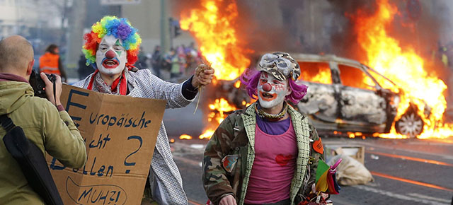Demonstrators dressed as clowns pass by a burning police car Wednesday, March 18, 2015 in Frankfurt, Germany. The Blockupy alliance said activists plan to try to blockade the new headquarters of the ECB to protest against government austerity and capitalism. (AP Photo/Michael Probst)