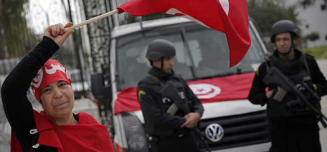 A woman waves the Tunisian flag as policemen stand guard at the National Bardo Museum two days after gunmen attacked the museum and killed scores of people in Tunis, Tunisia, Friday, March 20, 2015. The Islamic State group issued a statement Thursday claiming responsibility for the deadly attack on Tunisia's national museum that killed scores of people, mostly tourists. (AP Photo/Christophe Ena)