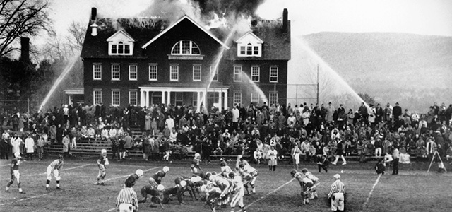 Spectators divide their attention as the Mount Hermon, Mass. High School football team hosts Deerfield Academy during a structure fire in the Mount Hermon science building Nov. 24, 1965. The science building was destroyed, and Mount Hermon lost the football game, ending a two-year-long winning streak. (AP Photo)