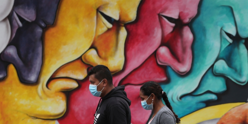 Un uomo e una donna, indossando maschere protettive come precauzione contro la diffusione del coronavirus, passano davanti a un murale a Quito, in Ecuador, sabato 28 marzo 2020 (AP Photo/Dolores Ochoa)