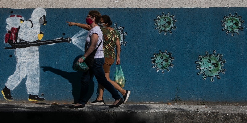Brasiliani che indossano maschere protettive passano davanti a un murale che raffigura un addetto alla sanificazione in equipaggiamento protettivo che spruzza detergente su un virus con la faccia del presidente Jair Bolsonaro nel quartiere di Estacio a Rio de Janeiro in Brasile, l'8 giugno 2020 (Bruna Prado/Getty Images)