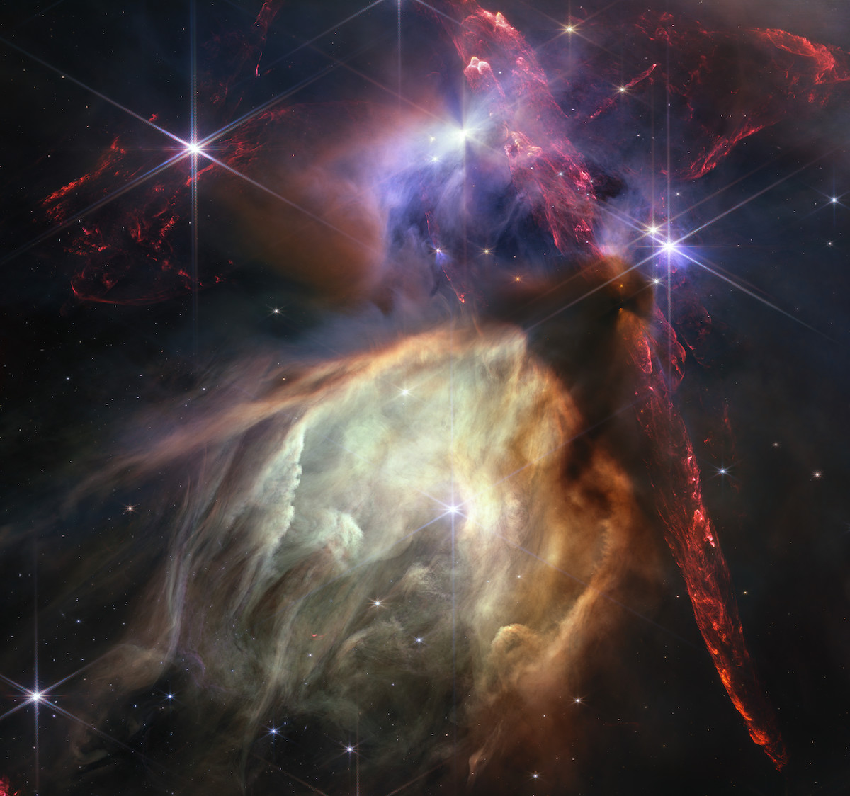 The first anniversary image from the NASA/ESA/CSA James Webb Space Telescope displays star birth like it’s never been seen before, full of detailed, impressionistic texture. The subject is the Rho Ophiuchi cloud complex, the closest star-forming region to Earth. It is a relatively small, quiet stellar nursery, but you’d never know it from Webb’s chaotic close-up. Jets bursting from young stars crisscross the image, impacting the surrounding interstellar gas and lighting up molecular hydrogen, shown in red. Some stars display the telltale shadow of a circumstellar disc, the makings of future planetary systems. The young stars at the centre of many of these discs are similar in mass to the Sun or smaller. The heftiest in this image is the star S1, which appears amid a glowing cave it is carving out with its stellar winds in the lower half of the image. The lighter-coloured gas surrounding S1 consists of polycyclic aromatic hydrocarbons, a family of carbon-based molecules that are among the most common compounds found in space. [Image description: Red dual opposing jets coming from young stars fill the darker top half of the image, while a glowing pale-yellow, cave-like structure is bottom centre, tilted toward two o’clock, with a bright star at its centre. The dust of the cave structure becomes wispy toward eight o’clock. Above the arched top of the dust cave three groupings of stars with diffraction spikes are arranged. A dark cloud sits at the top of the arch of the glowing dust cave, with one streamer curling down the right-hand side. The dark shadow of the cloud appears pinched in the centre, with light emerging in a triangle shape above and below the pinch, revealing the presence of a star inside the dark cloud. The image’s largest jets of red material emanate from within this dark cloud, thick and displaying structure like the rough face of a cliff, glowing brighter at the edges. At the top centre of the image, a star displays another, larger pinched dark shadow, this time vertically. To the left of this star is a more wispy, indistinct region.]