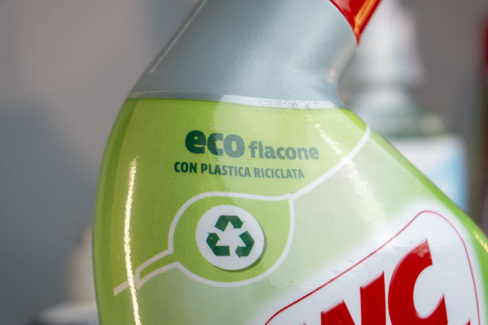 Dettaglio di un flacone di plastica grigia con un'etichetta coprente; l'etichetta segnala che è fatto di plastica riciclata