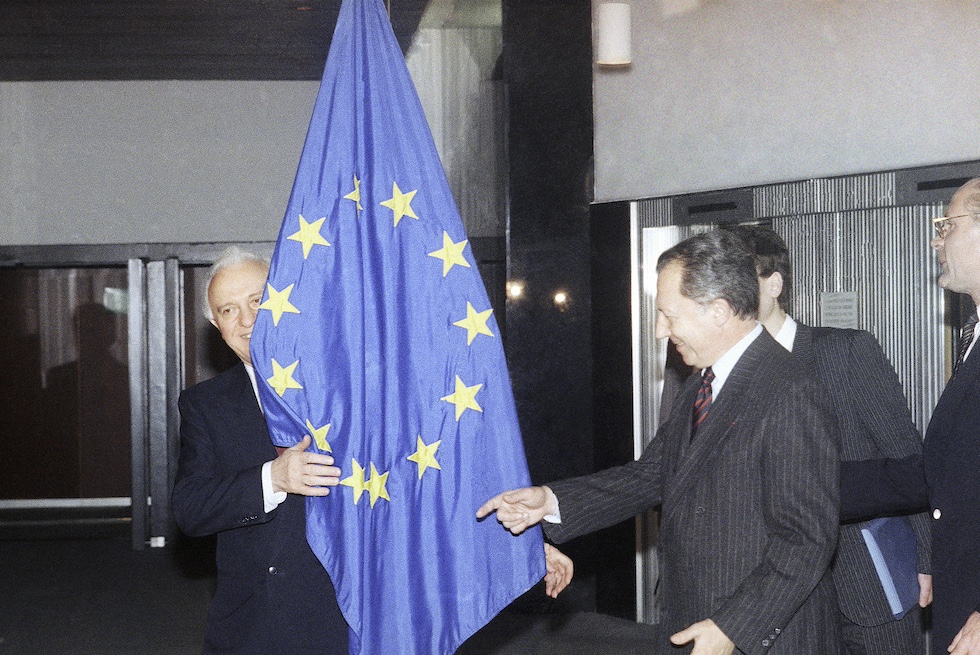Il ministro degli Esteri dell'Unione Sovietica Eduard Shevardnadze dietro alla bandiera della Comunità europea mentre il presidente della Commissione europea Jacques Delors gli indica il punto in cui dovrebbe trovarsi per la sessione fotografica nel 1989 (AP Photo/Carl Duyck)