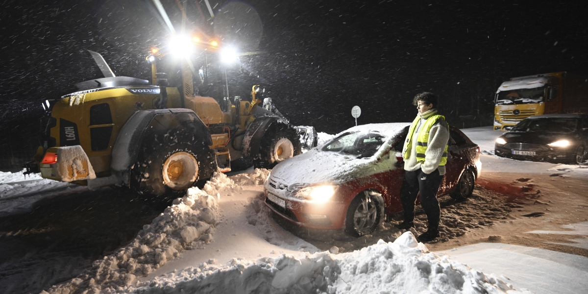una persona davanti a una macchina in diversi centimetri di neve, e dietro uno spazzaneve
