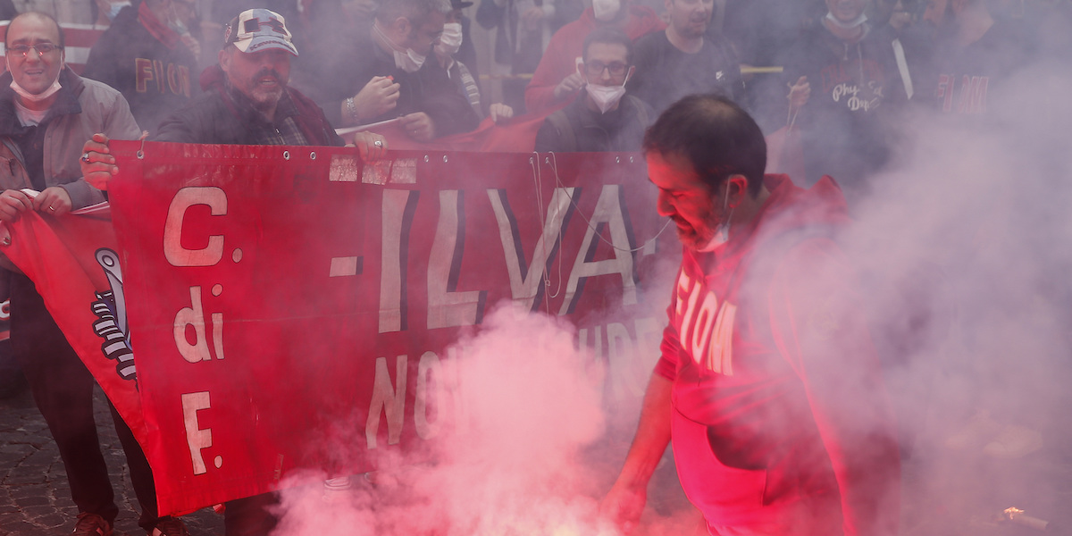 Una foto di una manifestazione dei sindacati con uno striscione rosso che dice "Ilva" e il fumo intorno