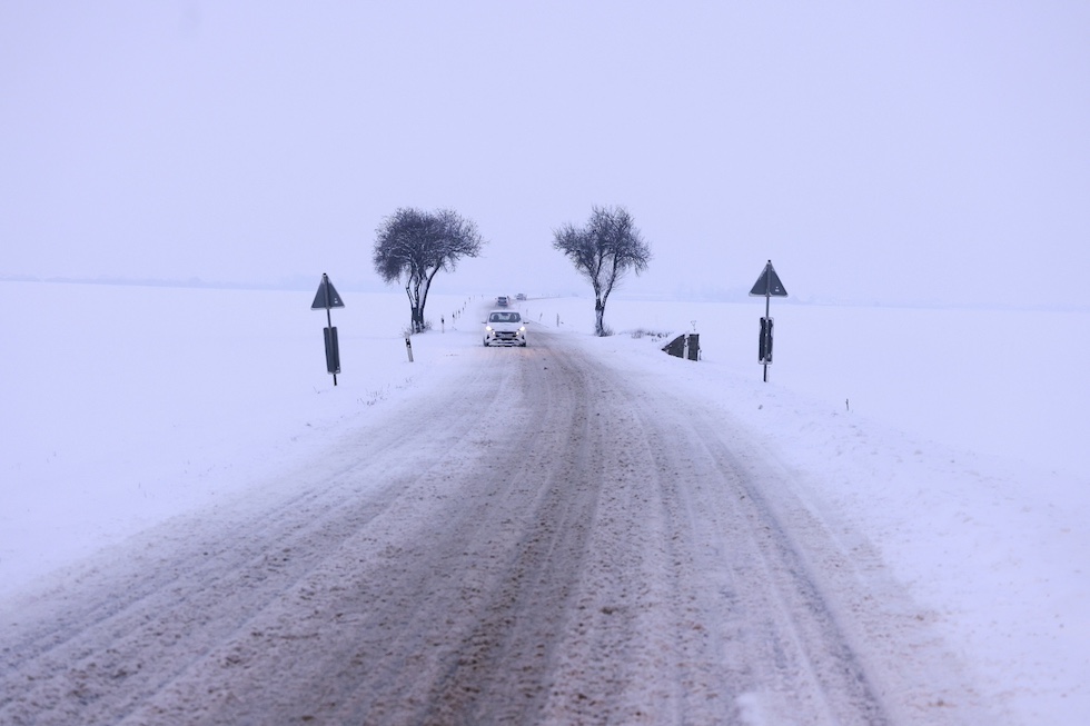 La foto mostra una macchina al centro di una strada di campagna bianca e un cielo bianco a causa della neve. La macchina sta passando fra due piccoli alberi neri.