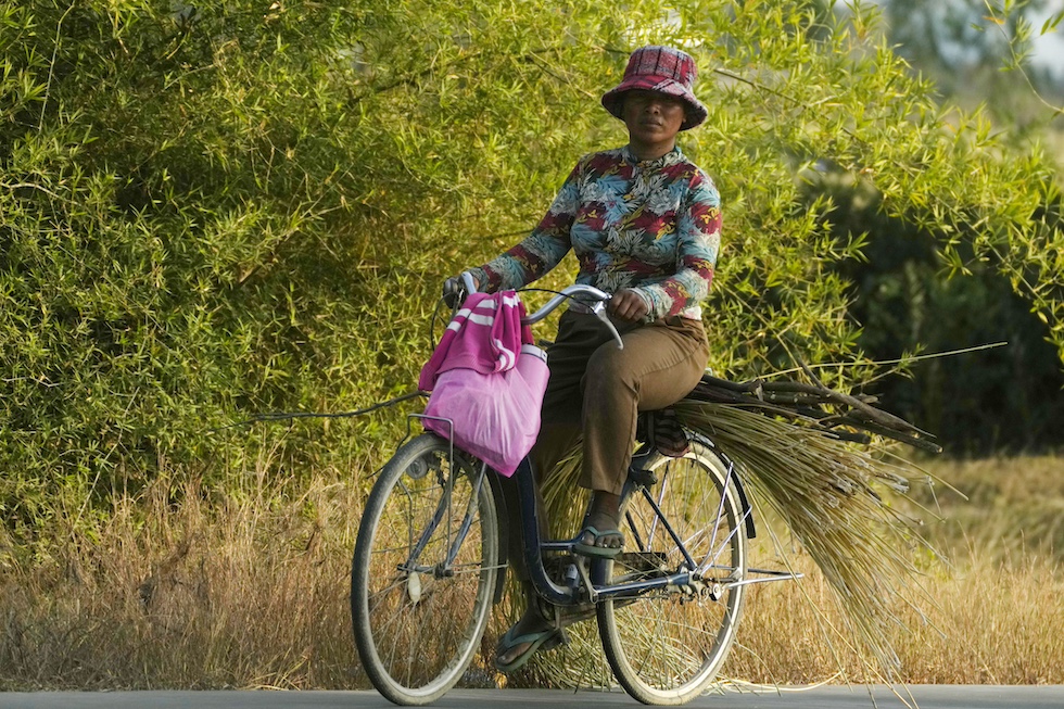 La foto mostra una donna in bicicletta in un paesaggio di campagna con dei bastoncini di legna sul portapacchi e una borsa rosa attaccata al manubrio nelle campagne fuori dalla capitale Phnom Penh