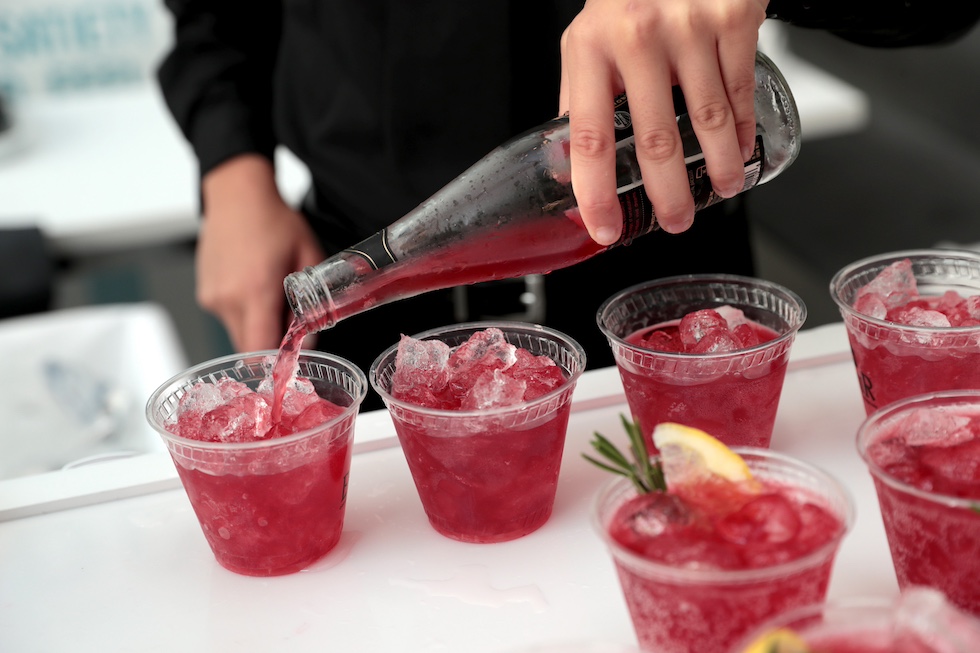 Un barista versa un cocktail analcolico rosso in sette bicchieri di plastica trasparente con ghiaccio, di cui uno con una scorza di limone e un rametto di rosmarino