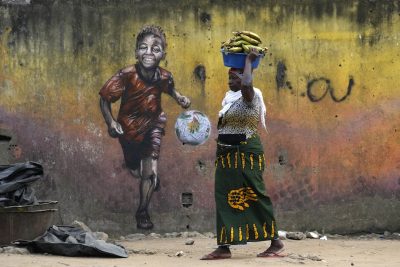 Una donna cammina davanti al murale di un bambino che gioca a calcio, portando un catino pieno di banane sulla testa. La foto è di martedì.