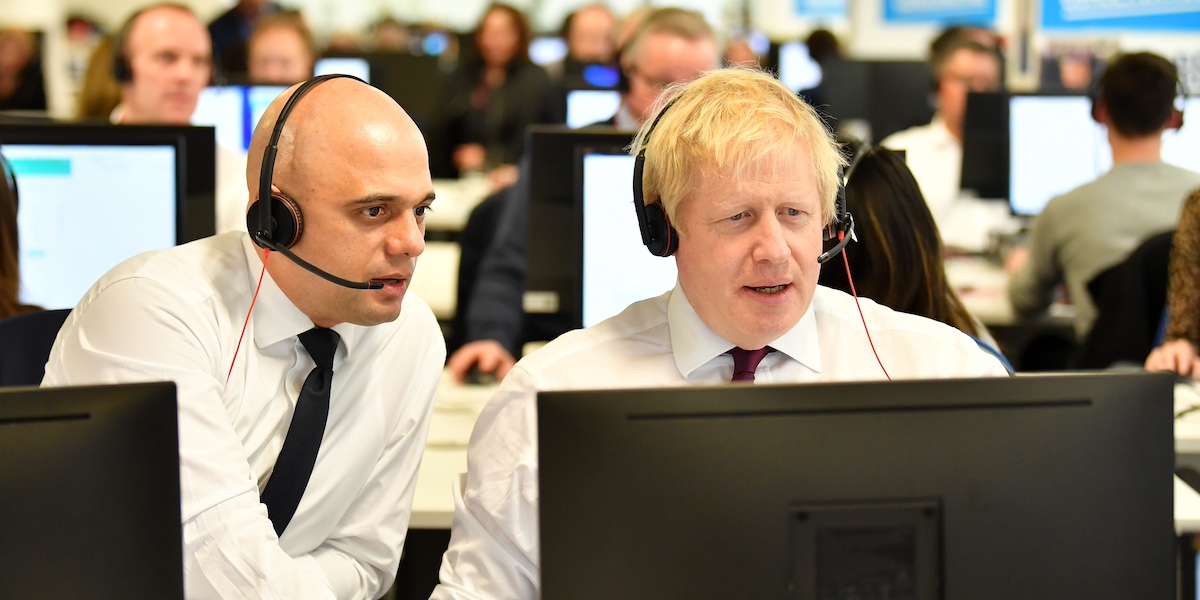 L’ex ministro dell’Economia del Regno Unito Sajid Javid e l’ex primo ministro Boris Johnson, seduti vicino, indossano cuffie con microfono del tipo utilizzato nei call center durante una telefonata nella sede del partito Conservatore