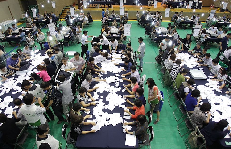Decine di scrutatori e scrutatrici seduti intorno a lunghi tavoli e impegnati nello scrutinio dei voti in una grande sala