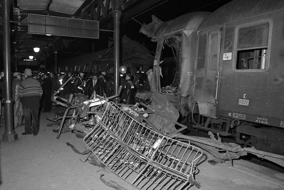 foto in bianco nere: le transenne e una carrozza del treno danneggiata dall'esplosione