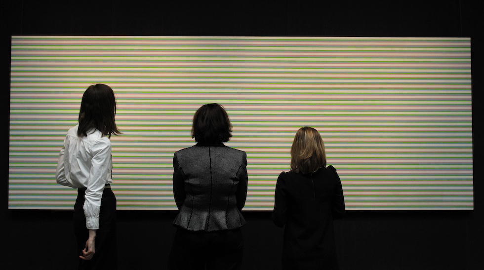 Tre persone osservano un dipinto composto da linee orizzontali, che formano un'illusione ottica