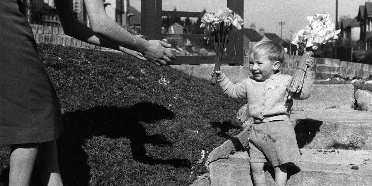 Un bambino piccolo con dei mazzi di fiori in mano e le mani della madre che si allungano verso di lui, in una fotografia degli anni Quaranta