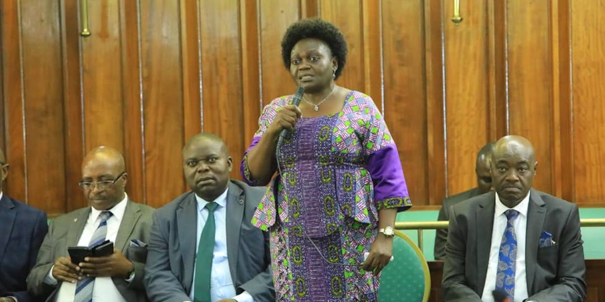 Una foto di Sarah Achieng Opendi mentre parla in parlamento in piedi con un vestito viola accanto a tre uomini seduti