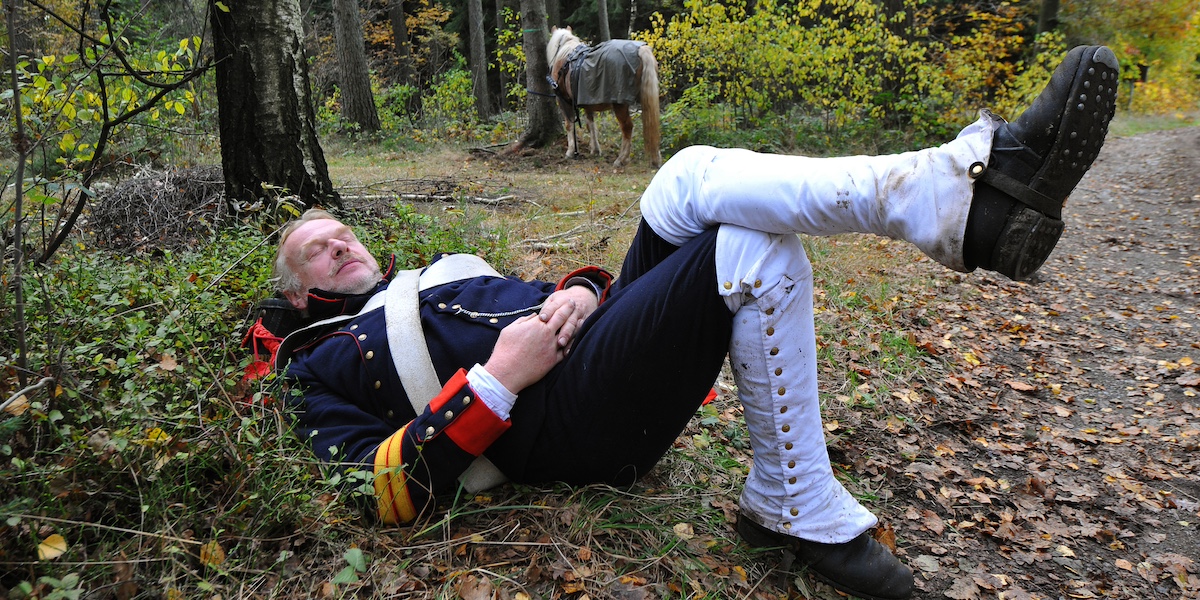 Un uomo vestito da soldato della fanteria napoleonica fa un pisolino sdraiato in un bosco, mentre sullo sfondo un cavallo è legato a un albero