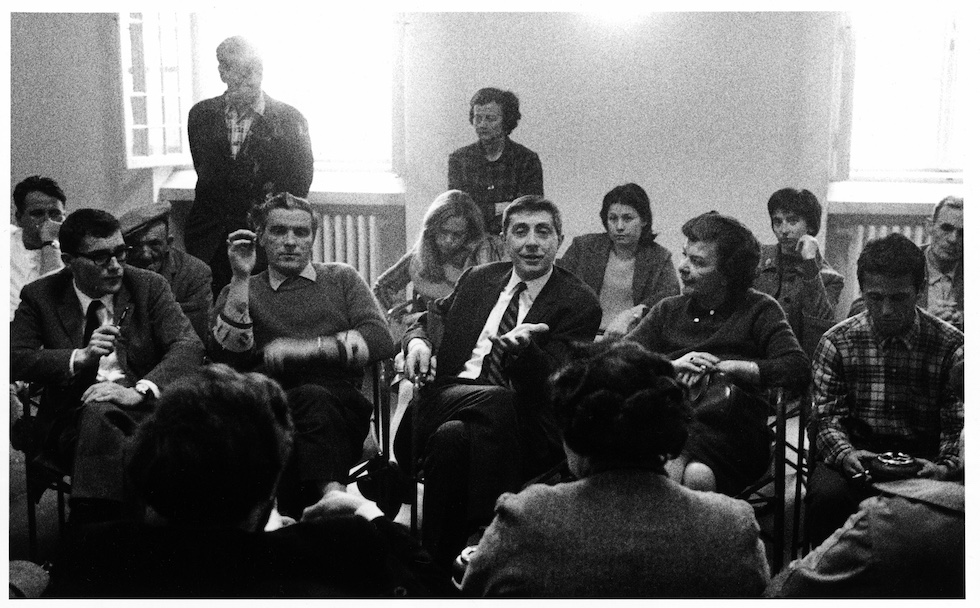 Basaglia discute durante una riunione, tra altre persone sedute vicino in cerchio una stanza affollata