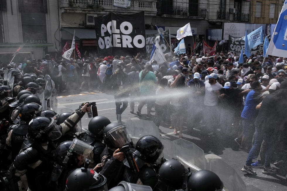 Due file di poliziotti sulla sinistra spruzzano gas lacrimogeno a dei manifestanti durante una protesta antigovernativa contro la scarsità di cibo nelle mense dei poveri e le riforme economiche proposte dal presidente Javier Milei