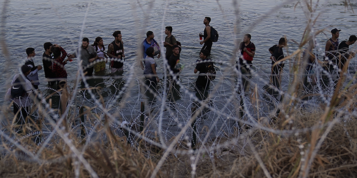 Un gruppo di migranti nelle acque del Rio Grande, visti attraverso una rete di filo spinato
