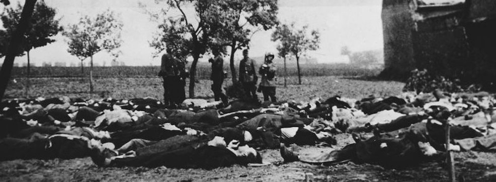 foto in bianco e nero con decine di uomini morti a terra e quattro persone in piedi dietro di loro, al centro della foto