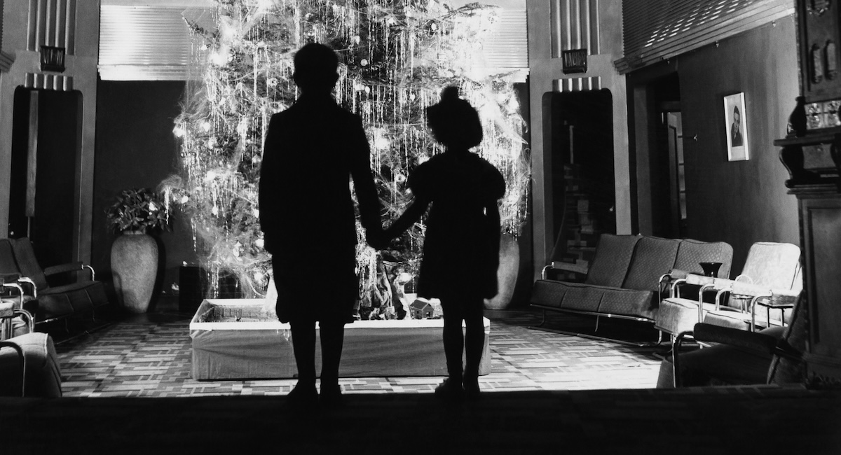 Due bambini si tengono per mano davanti a un albero di Natale senza regali sotto