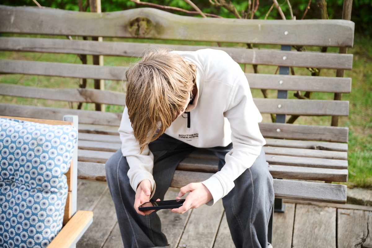 Un ragazzo guarda il suo smartphone mentre sta seduto su una panchina (Annette Riedl/dpa via ANSA)